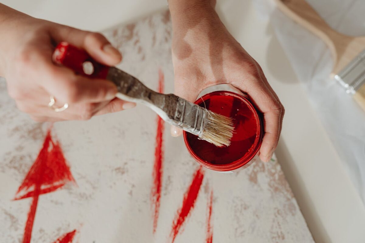 Close-up of hands dipping a paintbrush into red paint, creating abstract designs.